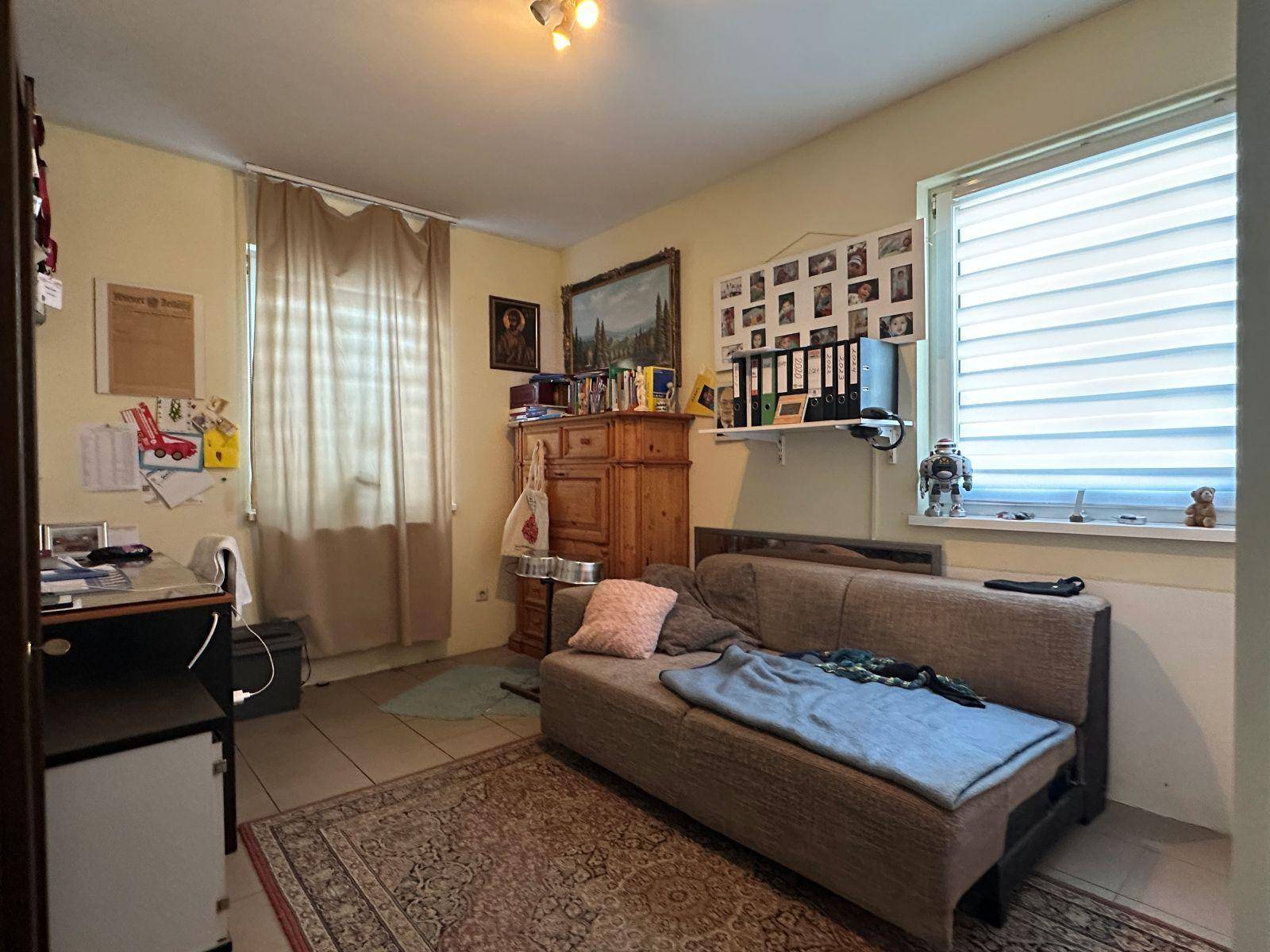Helles Zimmer mit Fliesenboden, Schlafsofa und Fenster mit Jalousien.