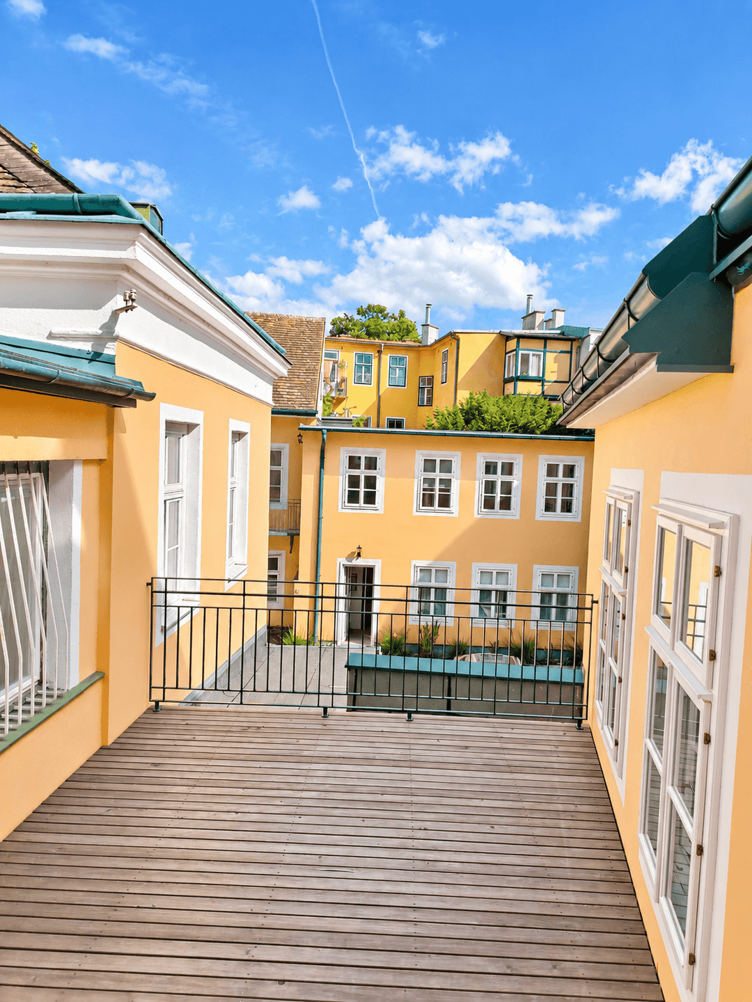 Holzterrasse mit Blick auf einen Innenhof und weitere gelbe Gebäude unter blauem Himmel.