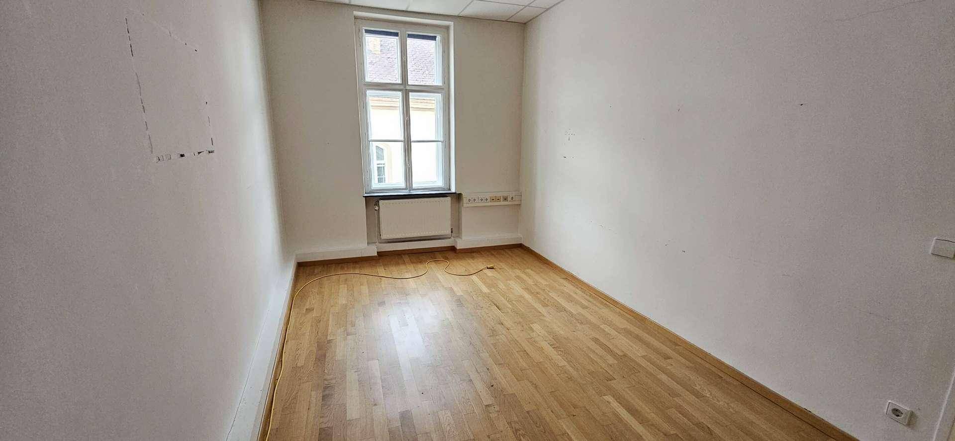 Kleinerer Büroraum mit Parkettboden, einem Fenster und weißen Wänden.
