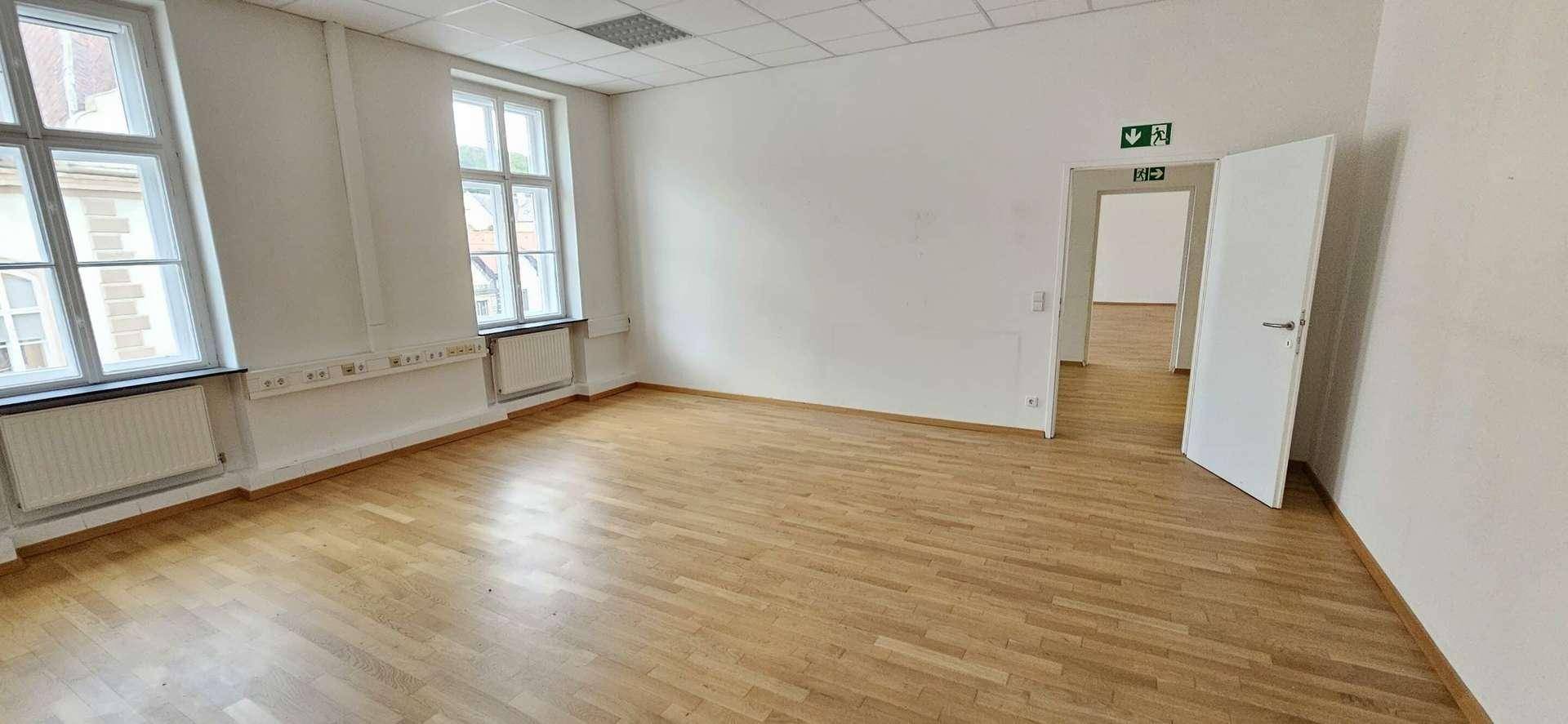 Helles, leeres Büro mit Parkettboden, großen Fenstern und weiß gestrichenen Wänden.