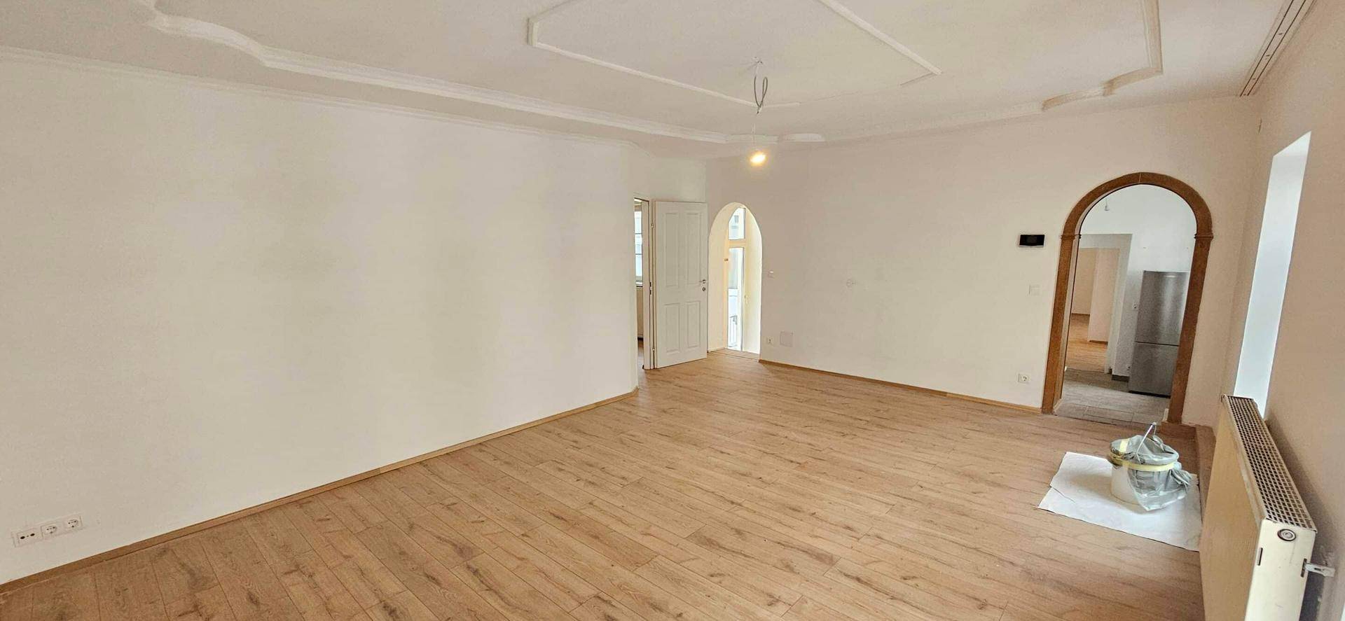 Großes, helles Zimmer mit Laminatboden, dekorativer Decke und Rundbogen zu einem weiteren Raum.