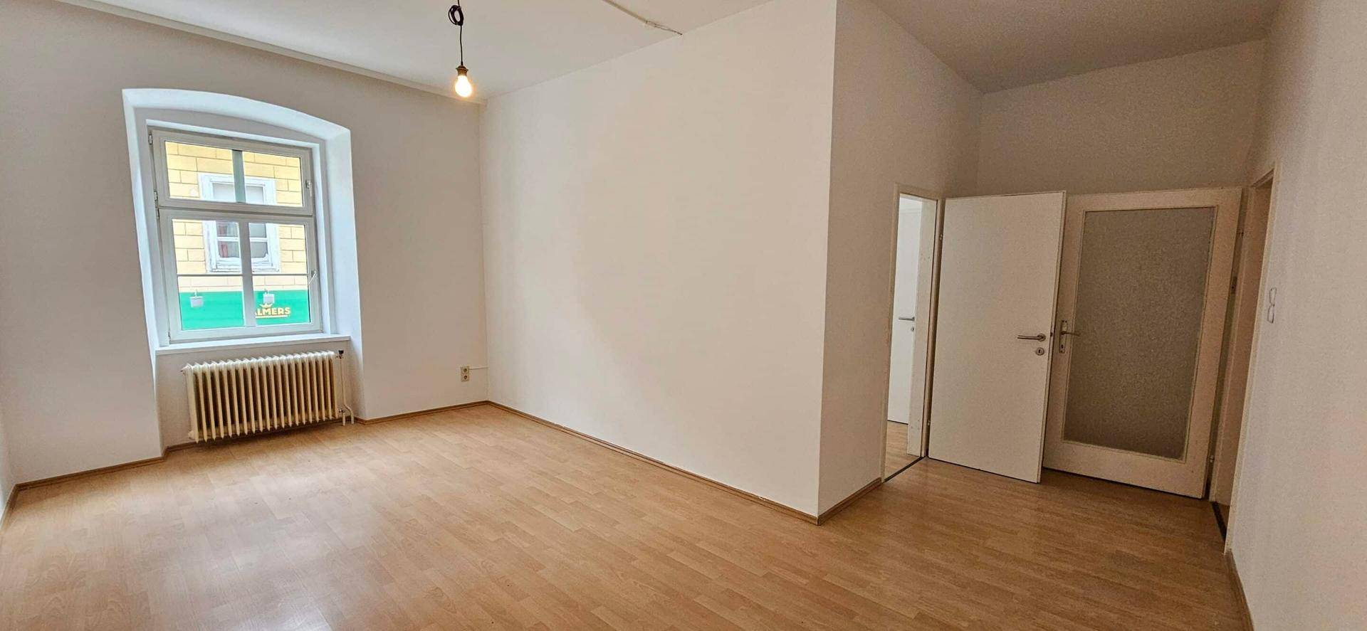 Helles, leeres Zimmer mit Laminatboden, einem großen Fenster und Heizkörper an der Wand.