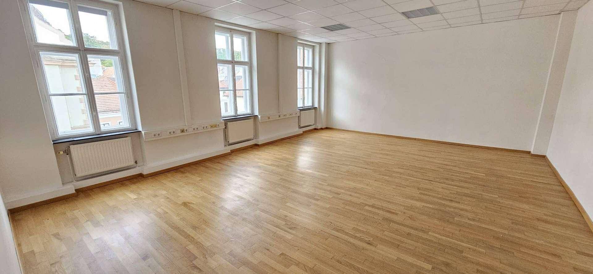 Leerer Büroraum mit Holzboden, drei großen Fenstern und weißer Decke.