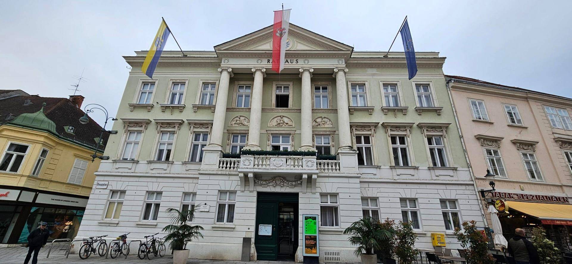 Repräsentatives historisches Gebäude mit Säulen, Balkon und Beflaggung unter bewölktem Himmel.