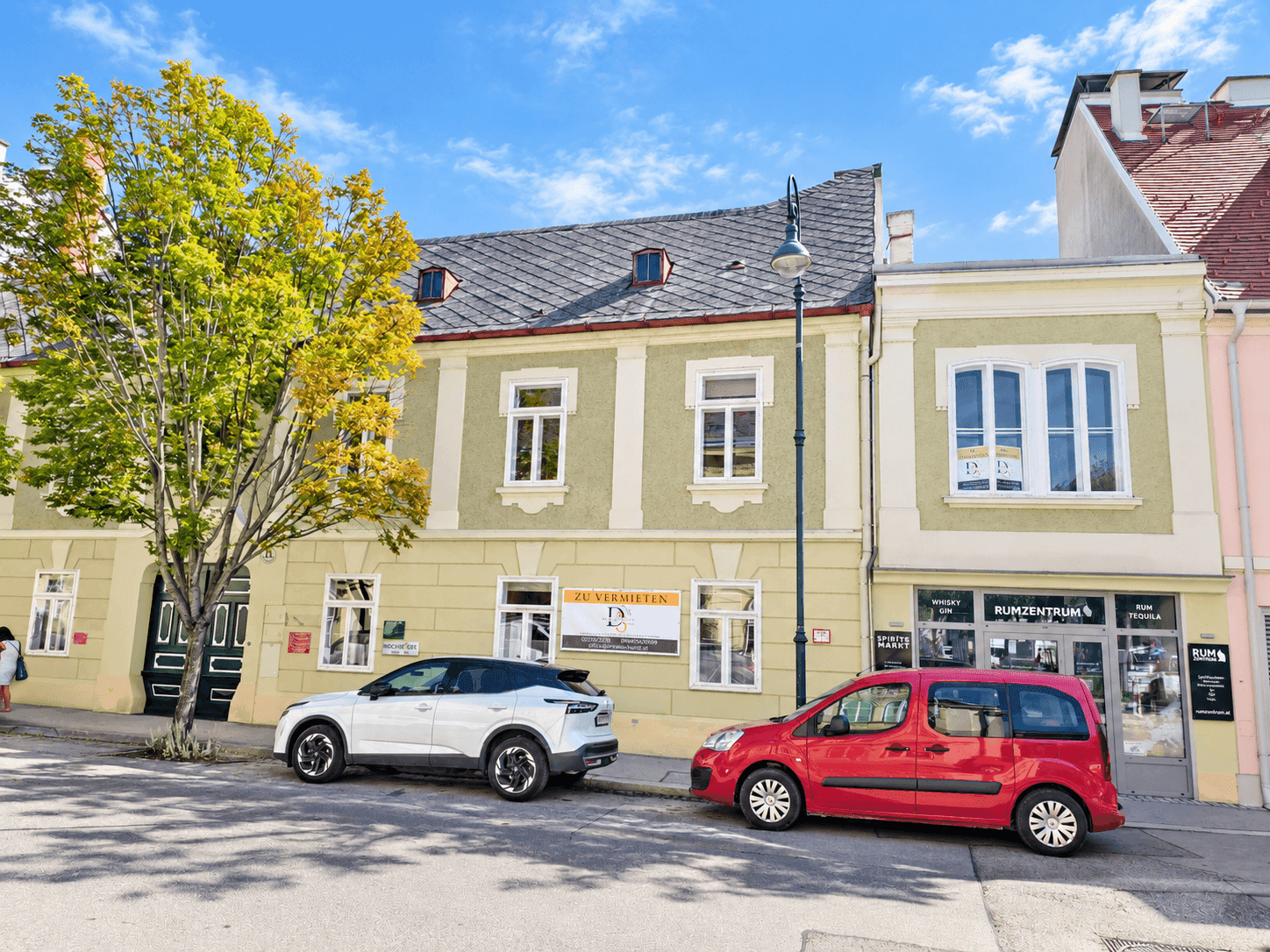 Straßenansicht mit geparkten Autos vor traditionellen Gebäuden, darunter ein Haus mit einem 'Zu Vermieten'-Schild.