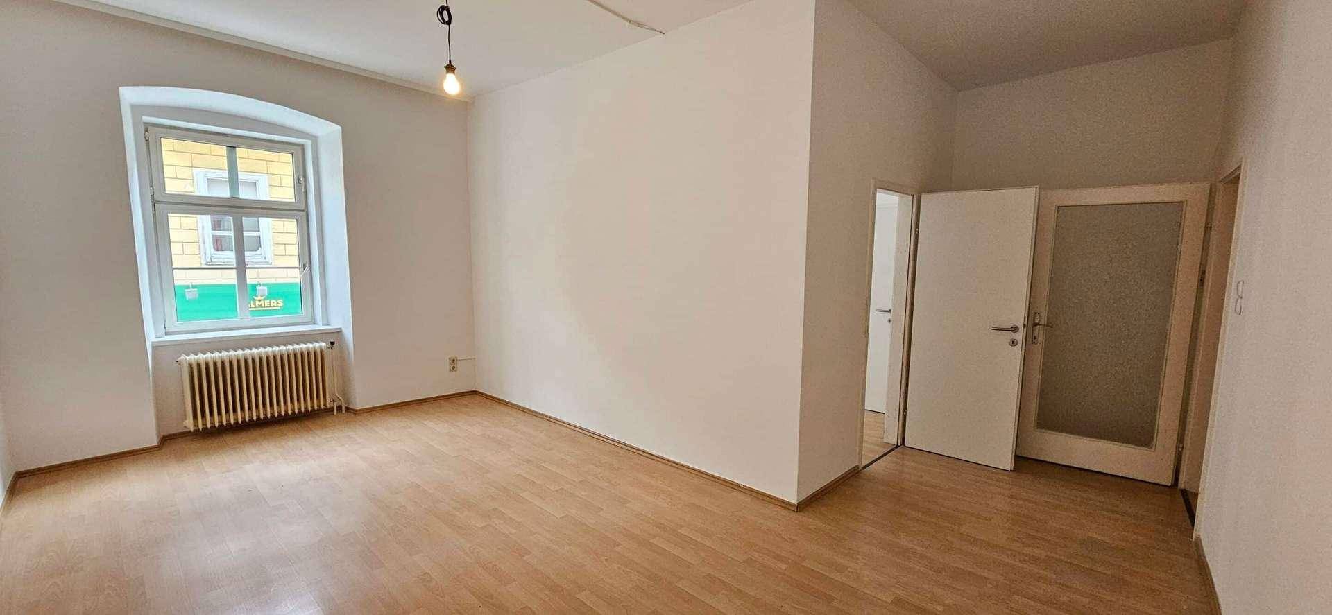Leeres Zimmer mit Holzboden, weißen Wänden, einem Fenster und einem Heizkörper.
