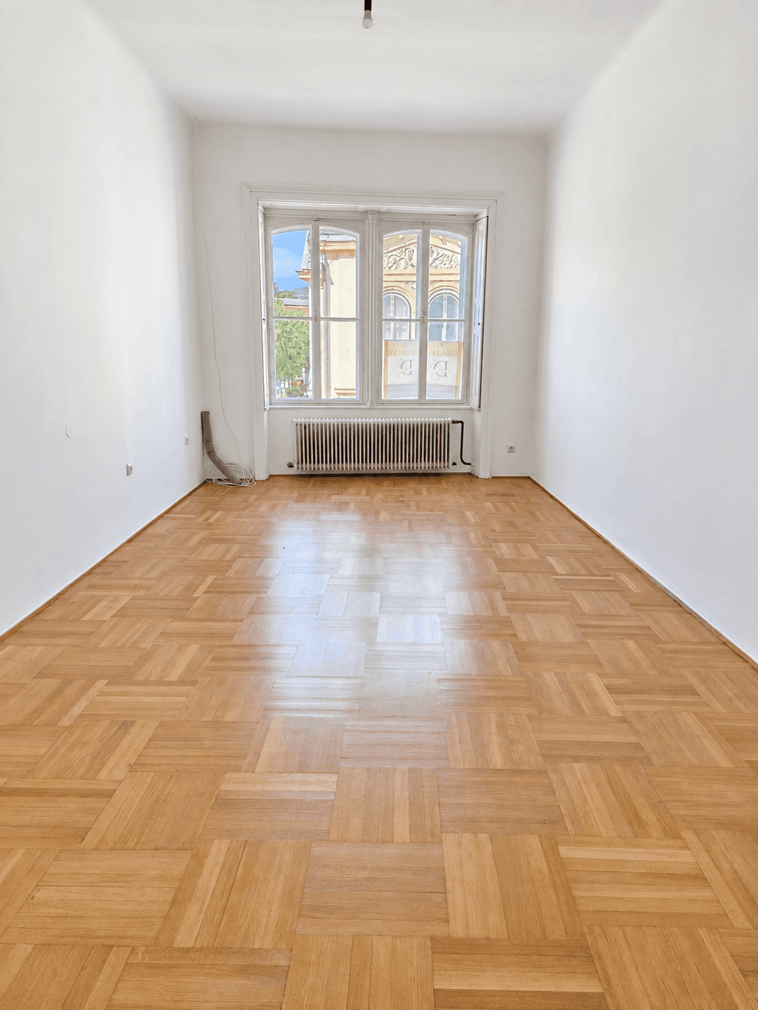 Leerer Raum mit Parkettboden und einem großen Fenster, das den Blick nach draußen freigibt.