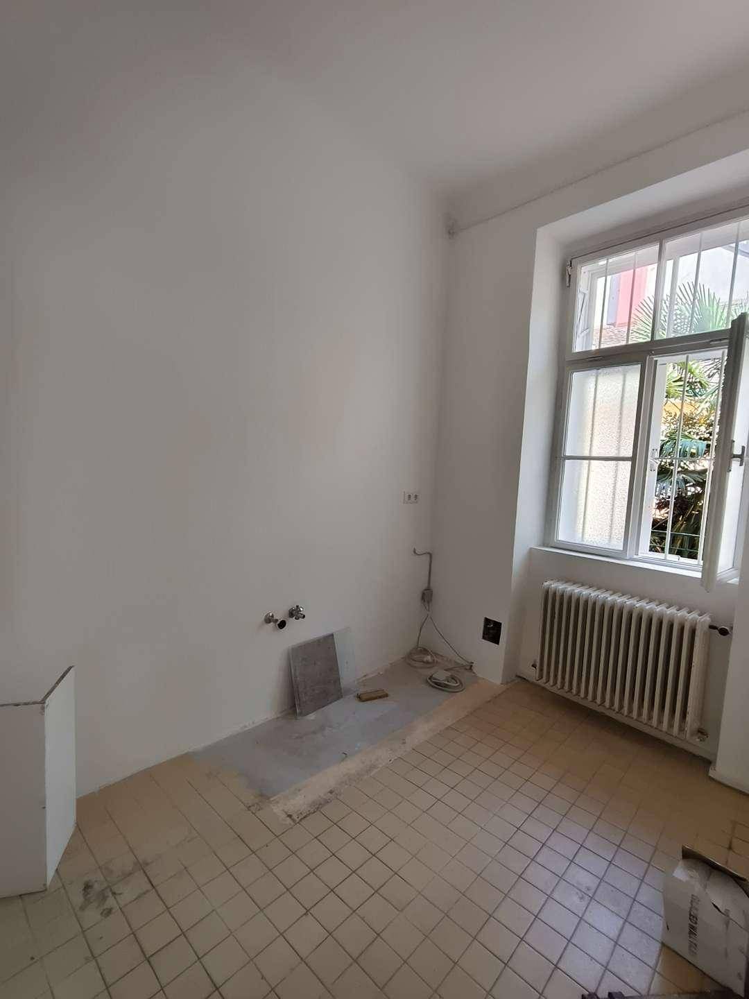 Leere Küche mit Fliesenboden, Heizkörper und einem Fenster mit Blick ins Grüne.
