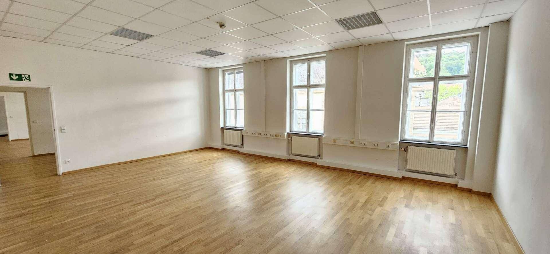 Geräumiges, leeres Büro mit Holzparkettboden, großen Fenstern und abgehängter Decke.