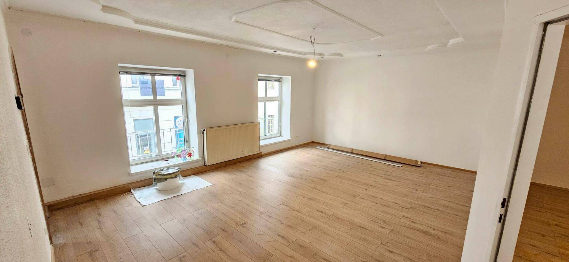 Geräumiges, helles Zimmer mit Laminatboden, zwei Fenstern und einer dekorativen Decke.