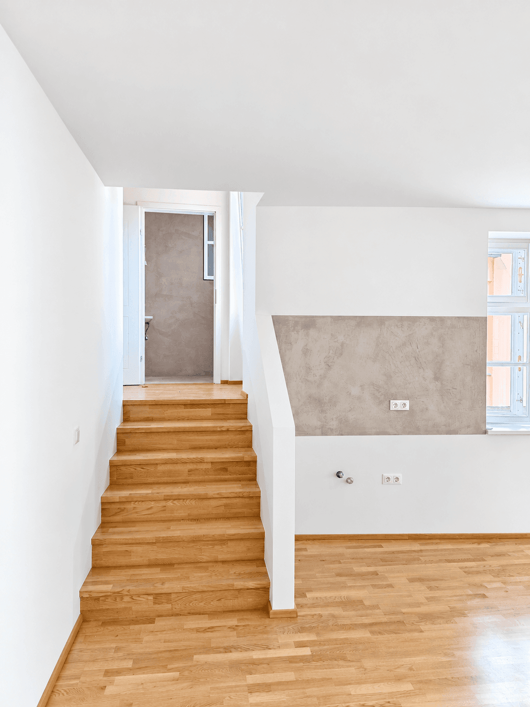 Holztreppe mit weißem Geländer in einem hellen Raum mit Holzboden und Fenstern.