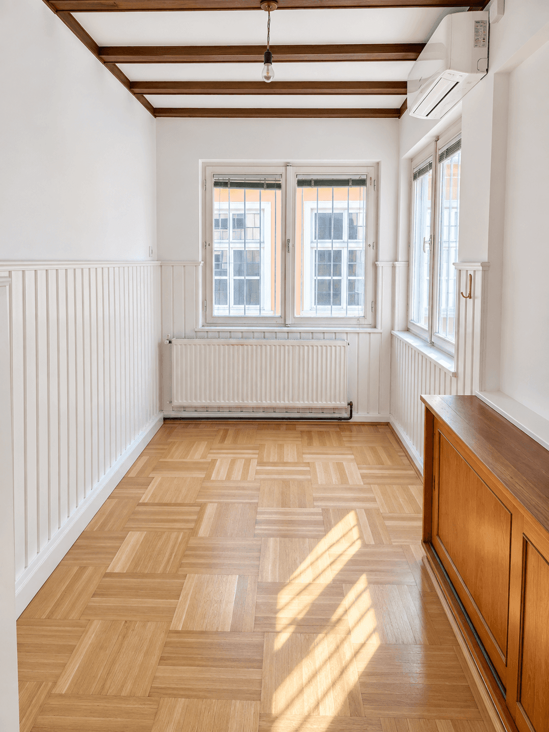 Schmaler Raum mit Holzbalken an der Decke, Holzvertäfelung und zwei Fenstern.