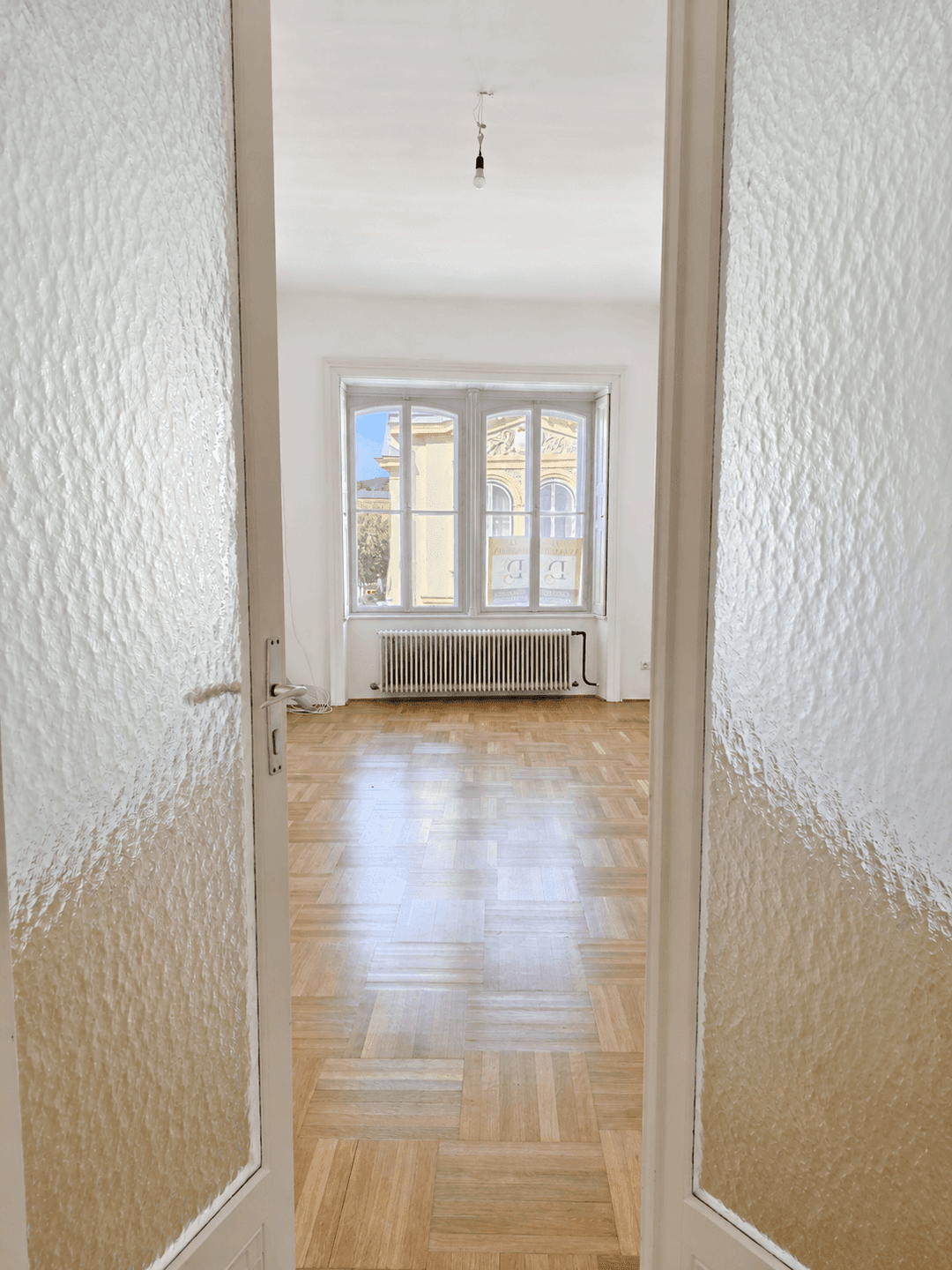 Blick durch eine Milchglastür in einen hellen Raum mit Parkettboden und großem Fenster.