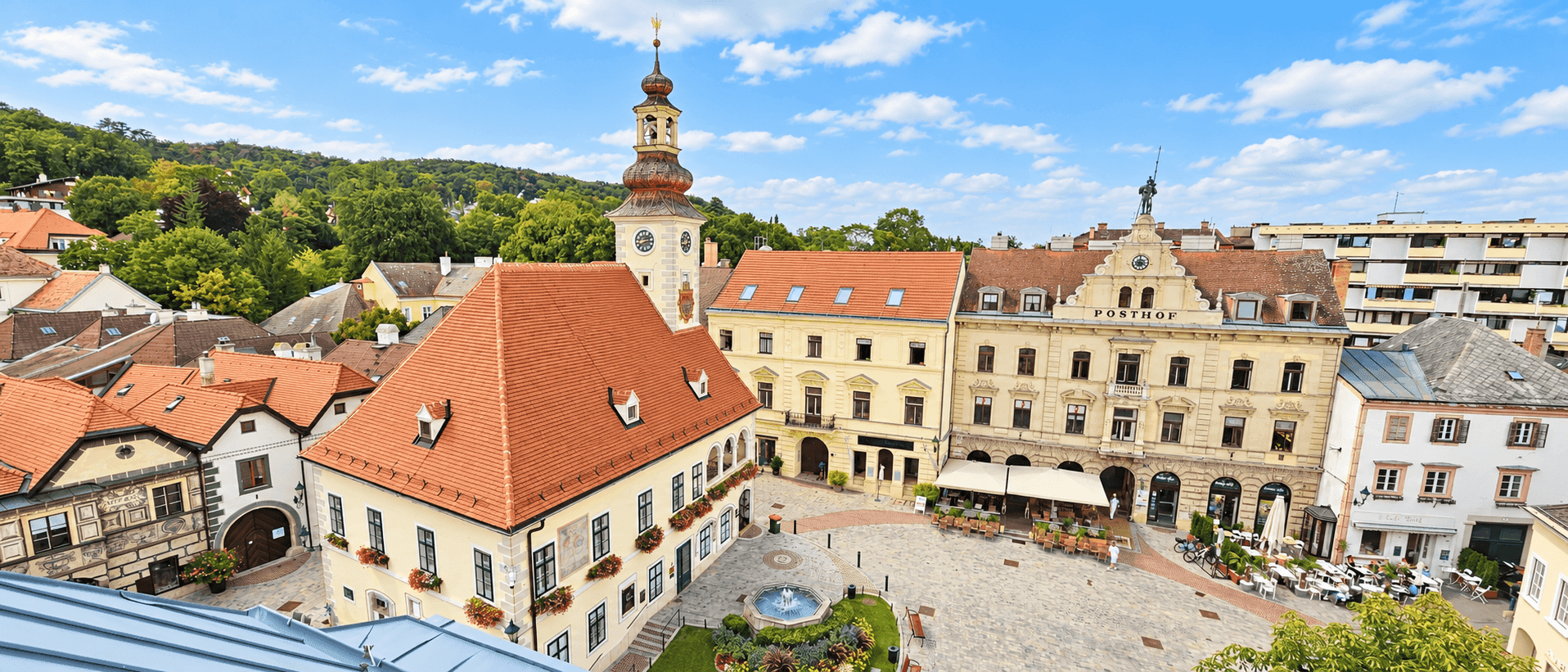 Panoramablick auf einen historischen Stadtplatz mit Brunnen, Kirchturm und traditionellen Gebäuden.