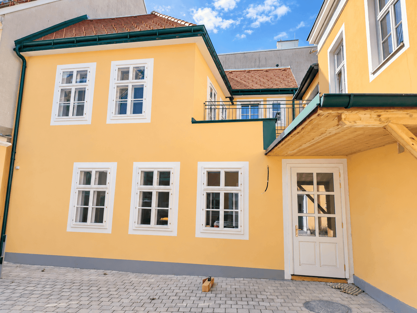 Gelbes Gebäude mit weißen Fenstern und einer gepflasterten Fläche davor.