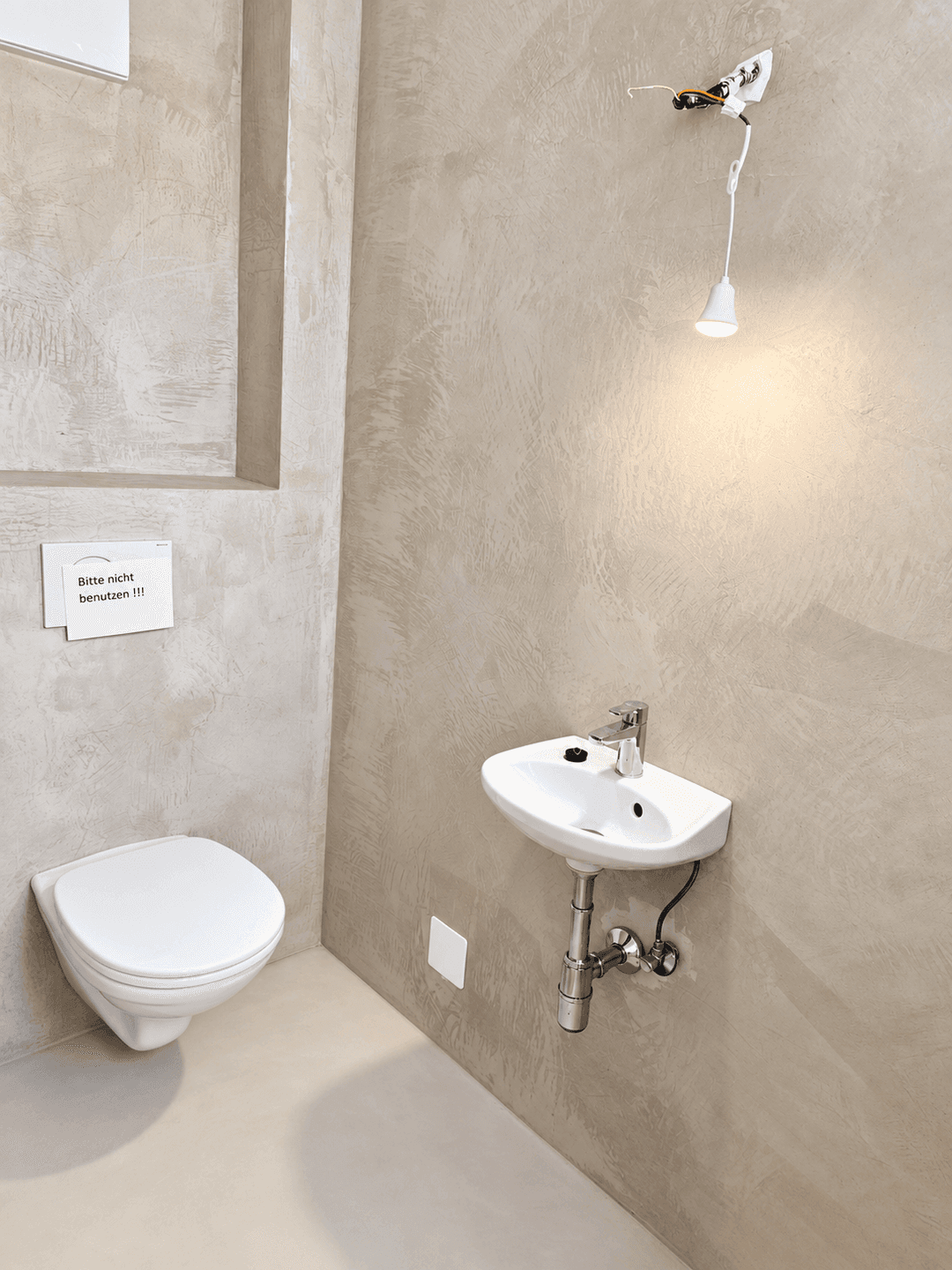 Gäste-WC mit Wand-WC, kleinem Waschbecken und provisorischer Beleuchtung.