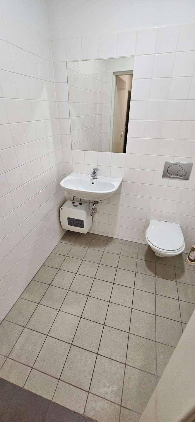 Einfache Toilette mit weißen Fliesen, Waschbecken, Spiegel und Wand-WC.