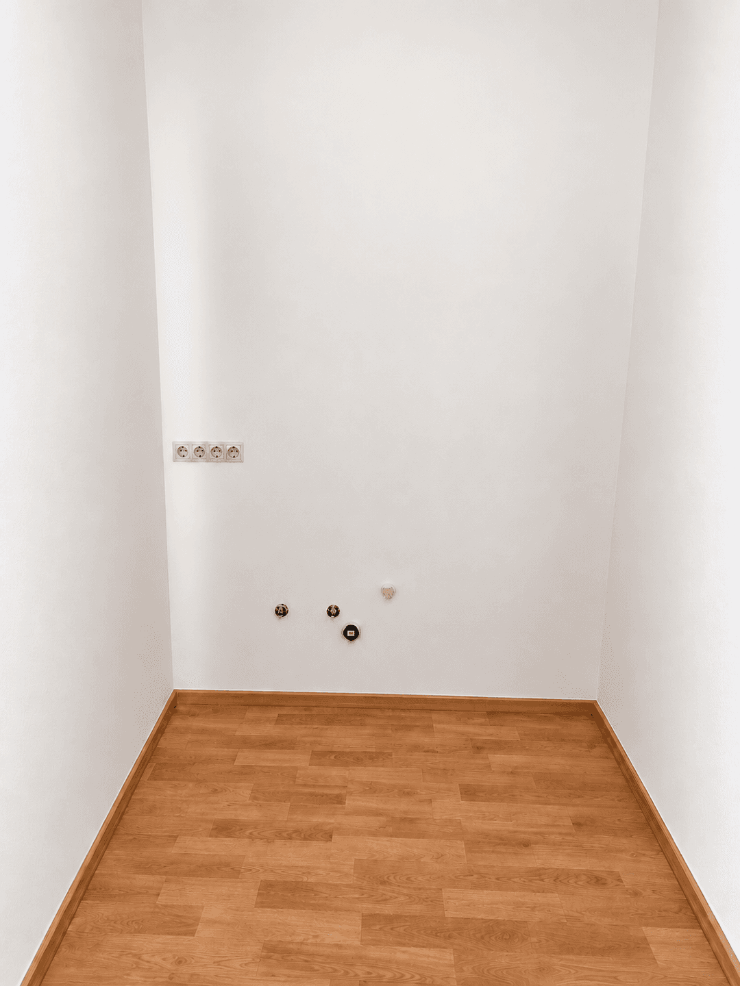 Kleiner, leerer Raum mit Laminatboden und Wandanschlüssen für Wasser und Strom.
