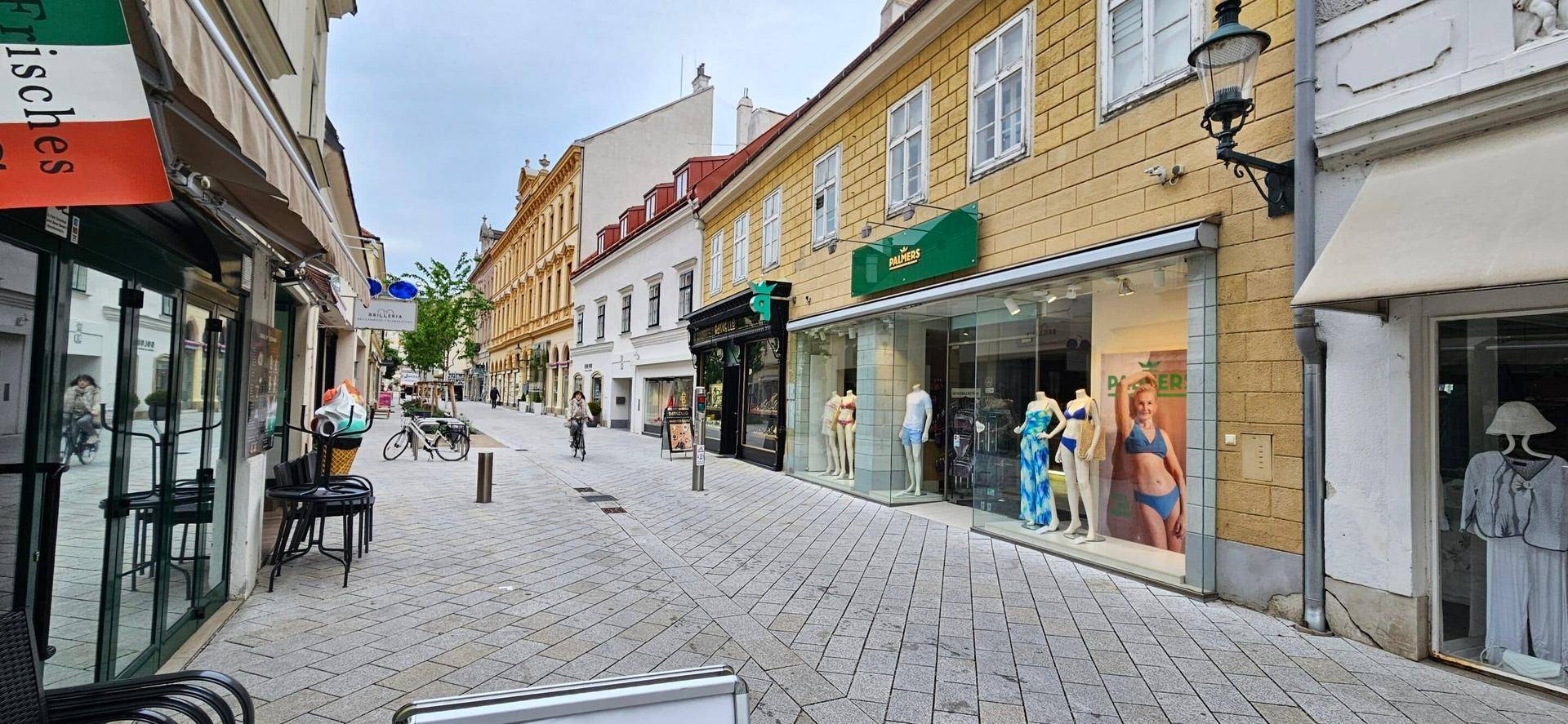 Belebte historische Gasse mit Kopfsteinpflaster, Geschäften und Schaufenstern unter bewölktem Himmel.