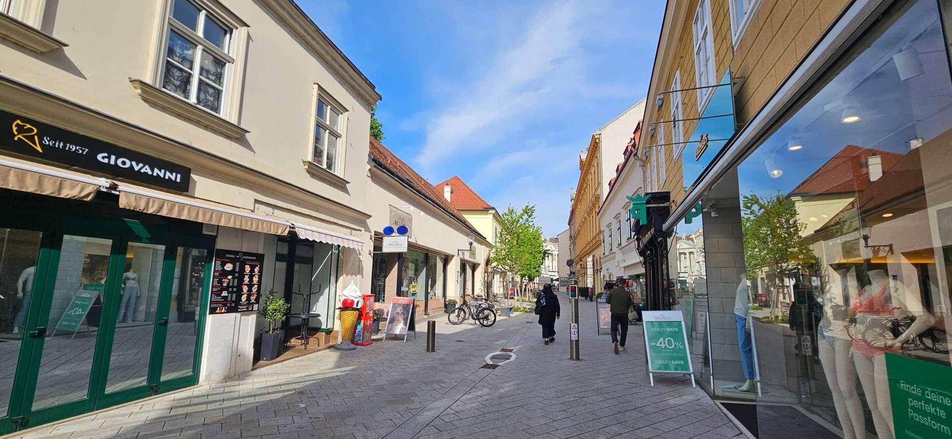 Belebte Fußgängerzone mit verschiedenen Geschäften und Passanten unter blauem Himmel.