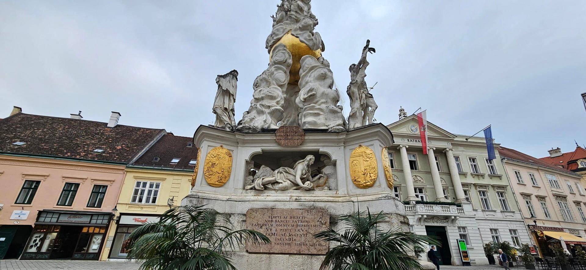 Historisches Denkmal auf einem Stadtplatz, umgeben von Gebäuden unter bewölktem Himmel.