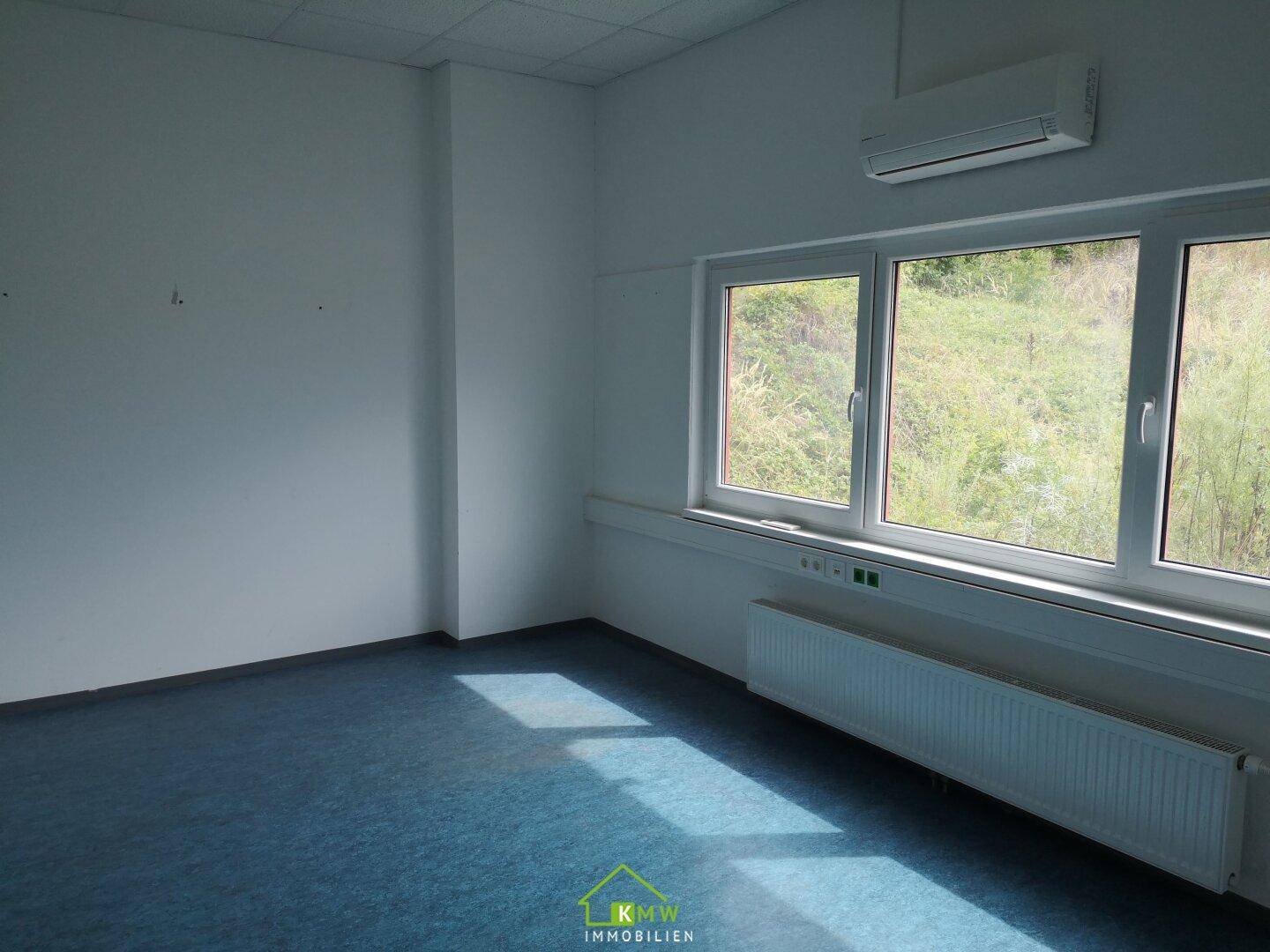 Leeres Büro mit blauem Teppichboden, großen Fenstern und einer Klimaanlage an der Wand.
