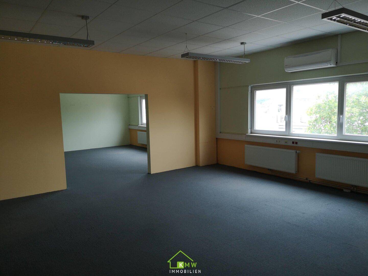 Leeres Büro mit dunkelgrauem Teppichboden, hellgelben Wänden und Klimaanlage, mit Blick ins Nebenzimmer.
