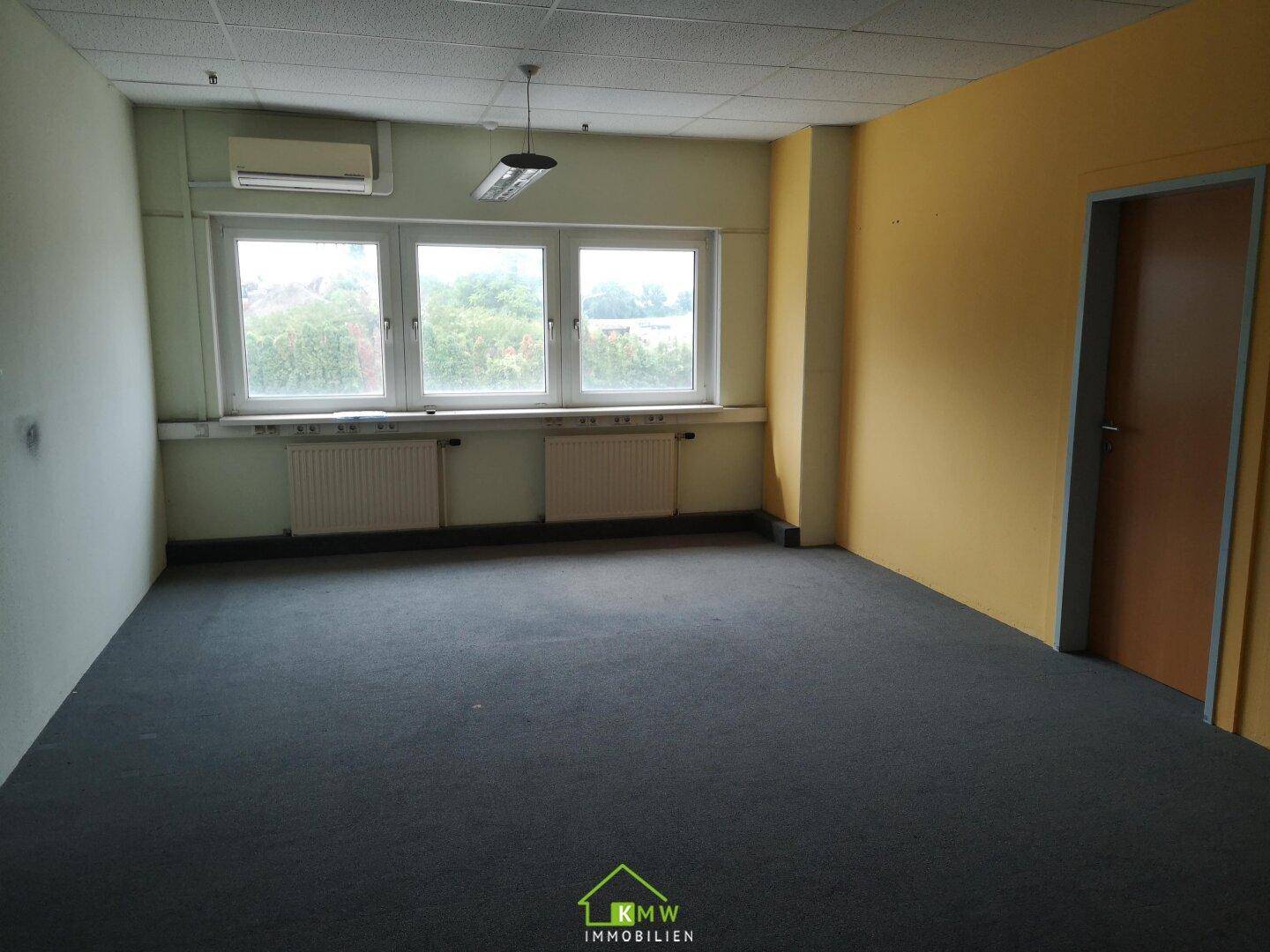 Unmöbliertes Büro mit grauem Teppichboden, Fenstern und einer gelben Akzentwand.