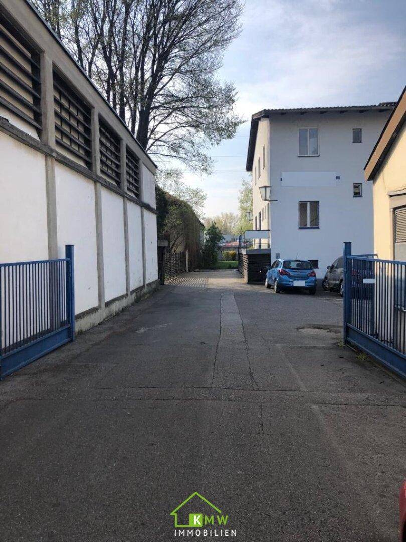 Einfahrt zwischen Gebäuden mit offenem blauen Tor und geparkten Autos unter hellem Himmel.
