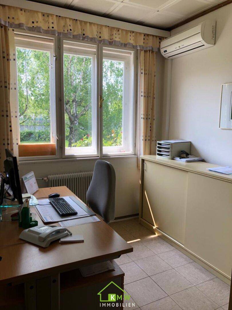 Helles Büro mit Schreibtisch, Stuhl und Blick ins Grüne durch die großen Fenster.
