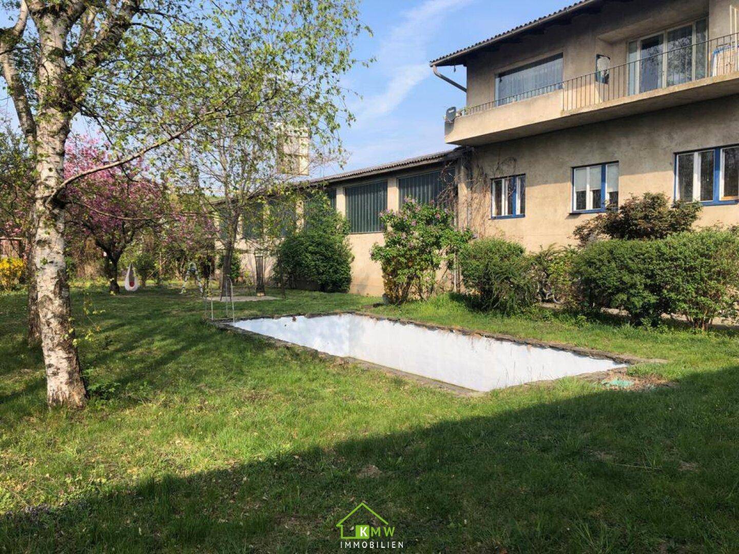 Großer Gartenbereich mit Bäumen, Rasen und einem leeren Schwimmbecken hinter dem Gebäude.