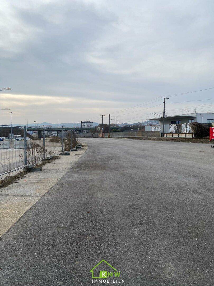 Blick über eine asphaltierte Fläche mit Bahngleisen und Gebäuden im Hintergrund unter bewölktem Himmel.