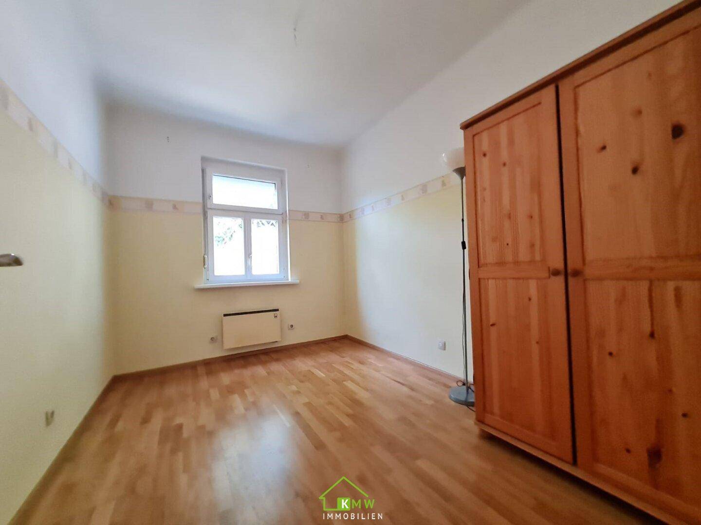 Schlafzimmer mit Holzfußboden, Fenster und einem großen Holzschrank.