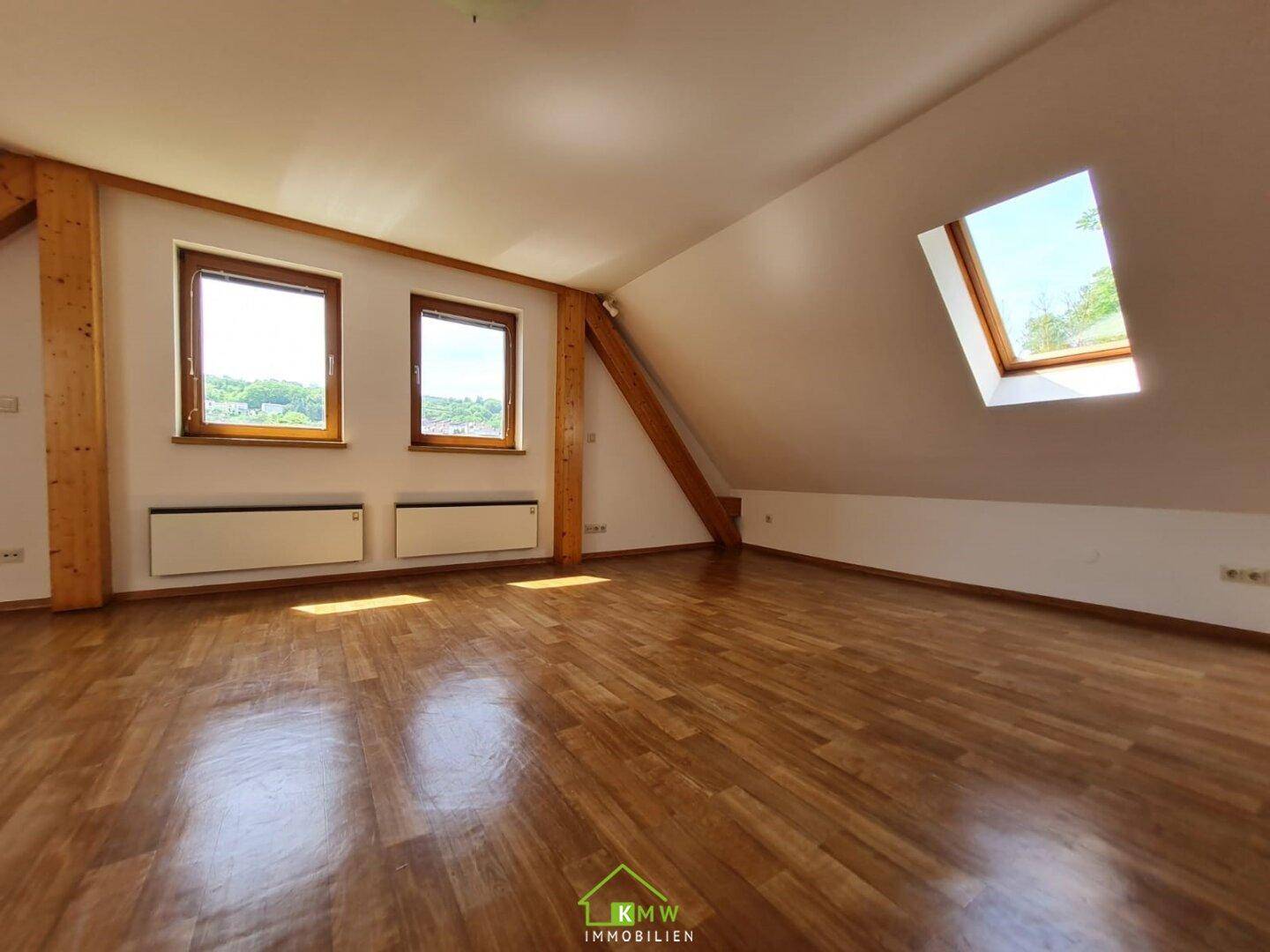Geräumiges Dachzimmer mit Holzfußboden, sichtbaren Holzbalken und mehreren Fenstern.