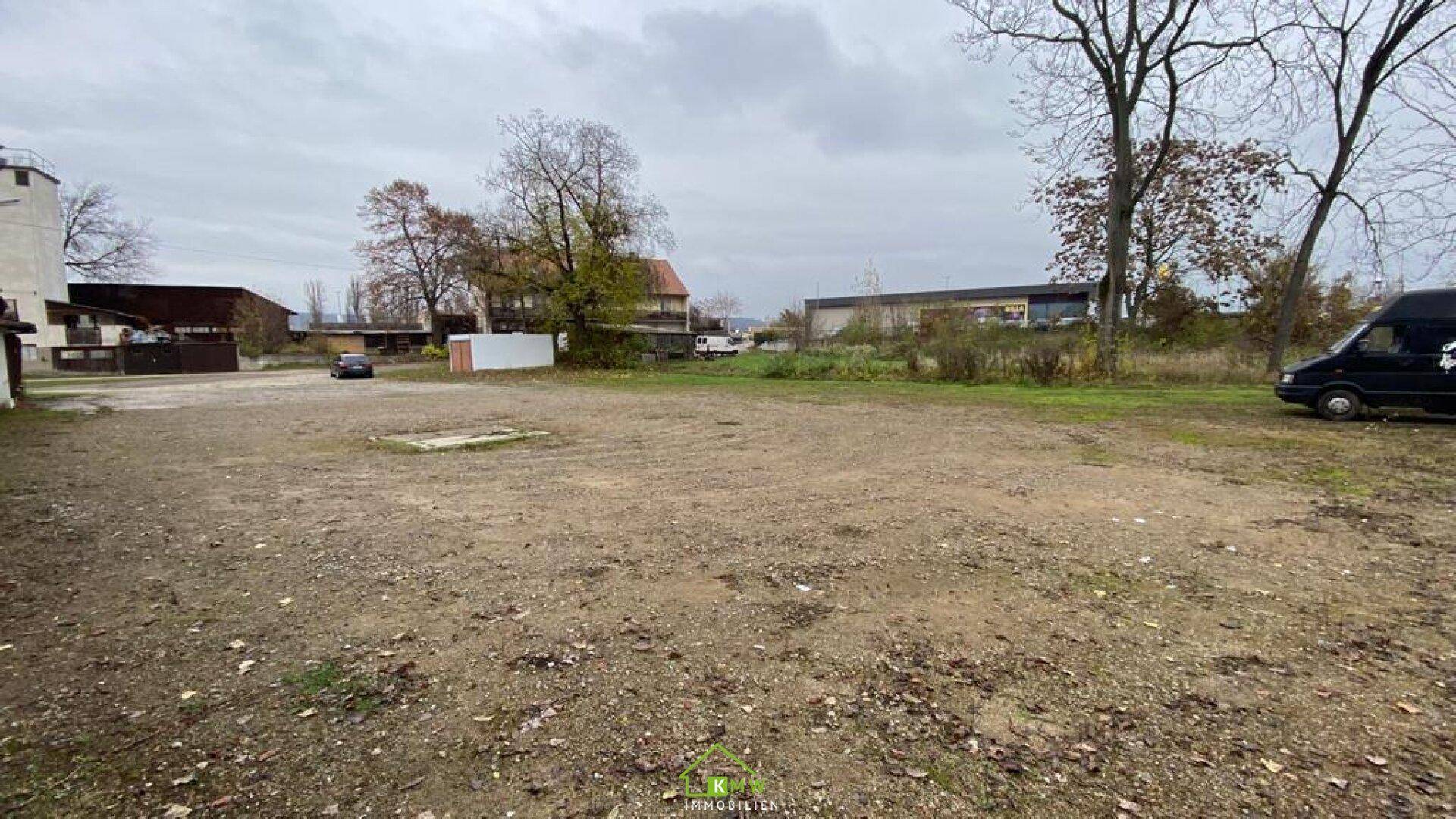 Weitläufiger, unbefestigter Außenbereich mit Bäumen und Gebäuden im Hintergrund unter bewölktem Himmel.