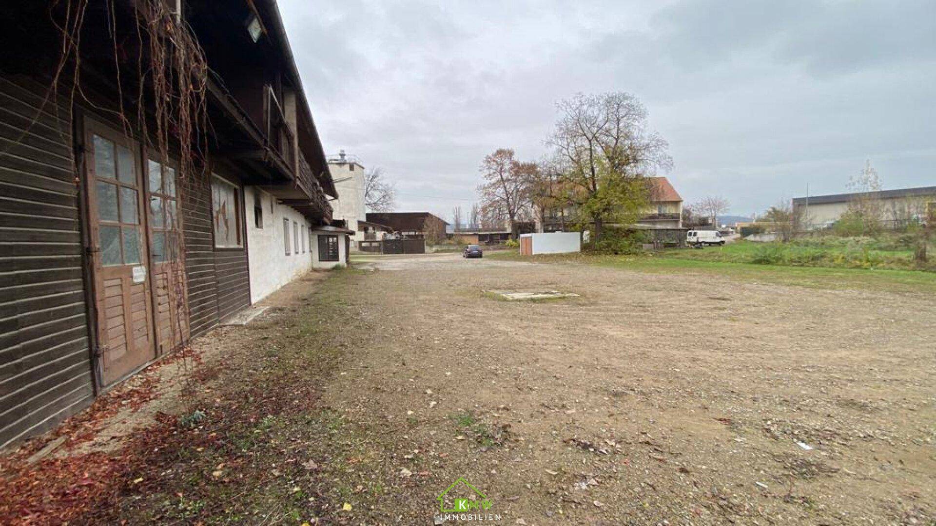 Weitläufiger, unbefestigter Außenbereich neben einem älteren Gebäude mit großen Holztüren, das Renovierung benötigt.
