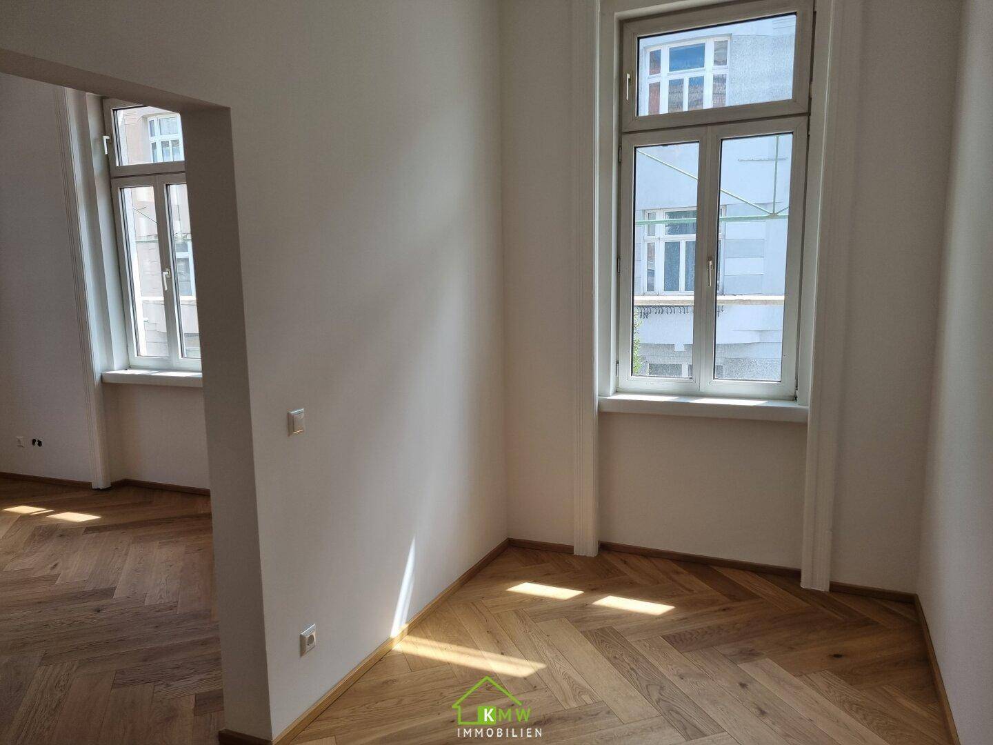 Lichtdurchfluteter Raum mit Fischgrätparkett und zwei Fenstern, die viel Helligkeit spenden.