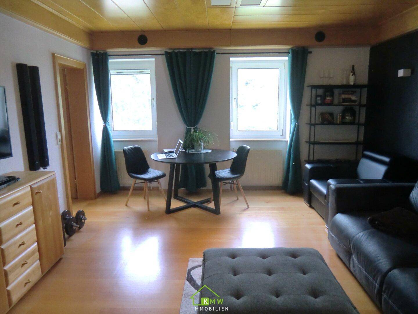 Geräumiges Wohnzimmer mit Holzboden, dunklem Sofa und zwei Fenstern mit grünen Vorhängen.