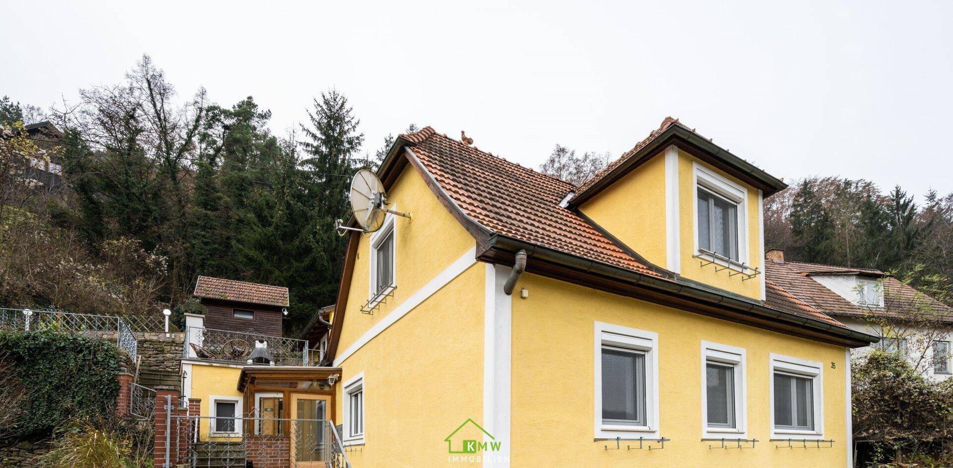 Seitenansicht des gelben Hauses mit rotem Ziegeldach und Fenstern mit Rollläden.