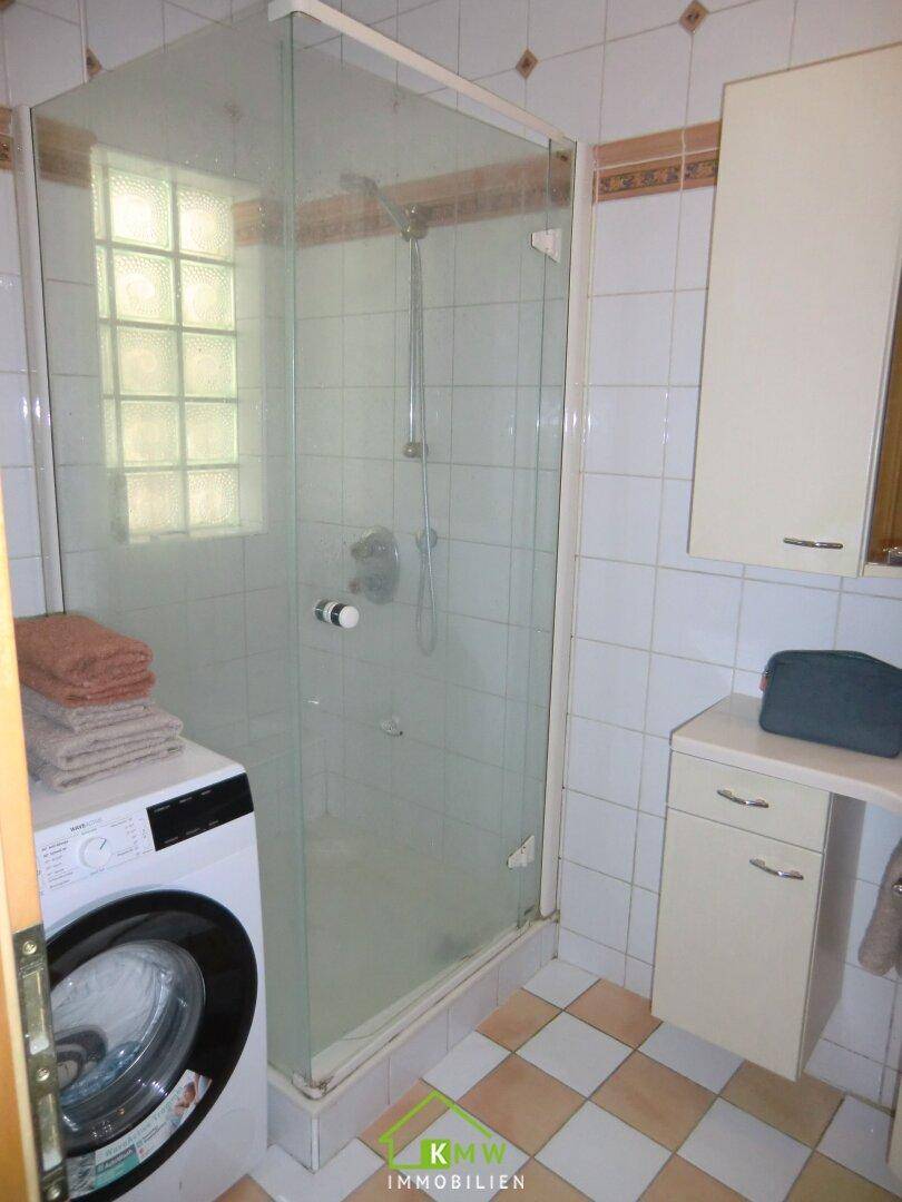 Badezimmer mit Duschkabine, Waschmaschine und hellen Fliesen im Schachbrettmuster.