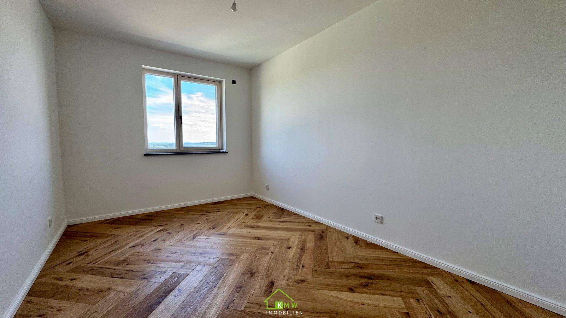 Leeres Schlafzimmer mit hellem Fischgrätparkett und einem Fenster, das viel Tageslicht hereinlässt.