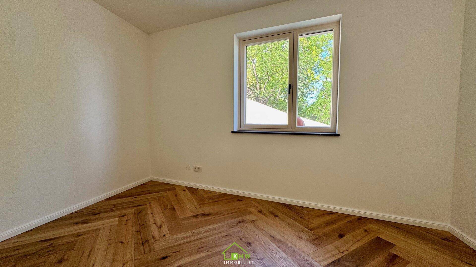 Helles Zimmer mit Fischgrätparkett und einem Fenster, das einen Blick ins Grüne bietet.