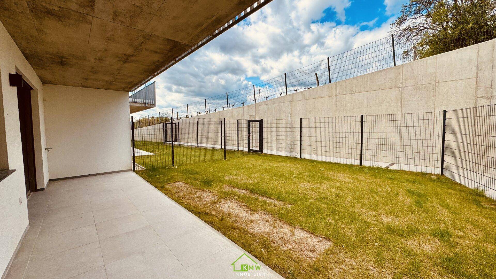 Private Terrasse mit angrenzendem Gartenbereich, umgeben von einem hohen Zaun und Betonwand.