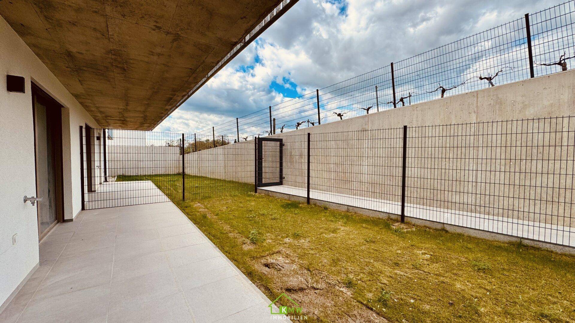 Gepflasterte Terrasse mit angrenzendem Rasenstreifen und hoher Umzäunung eines Neubaus.