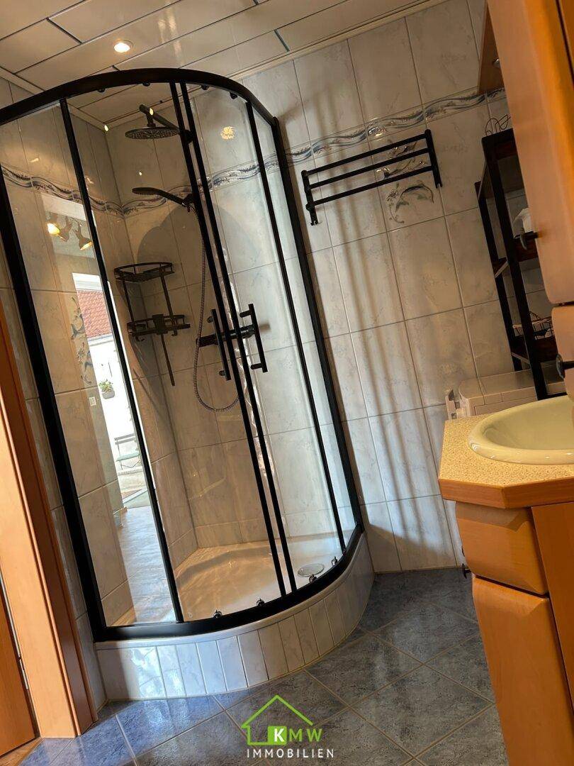 Badezimmer mit Eckduschkabine, hellen Fliesen und einem Waschbecken mit Unterschrank.