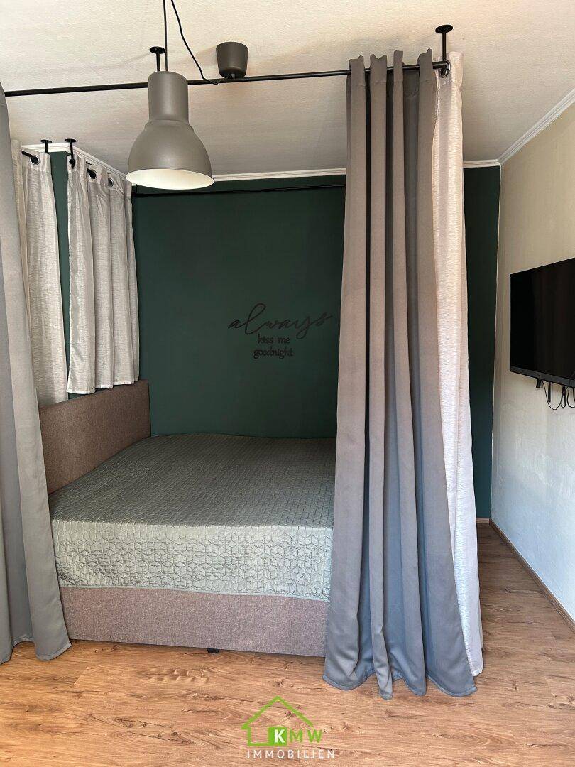 Schlafbereich mit Bett, dunkelgrüner Akzentwand und Vorhängen, auf einem Holzboden.