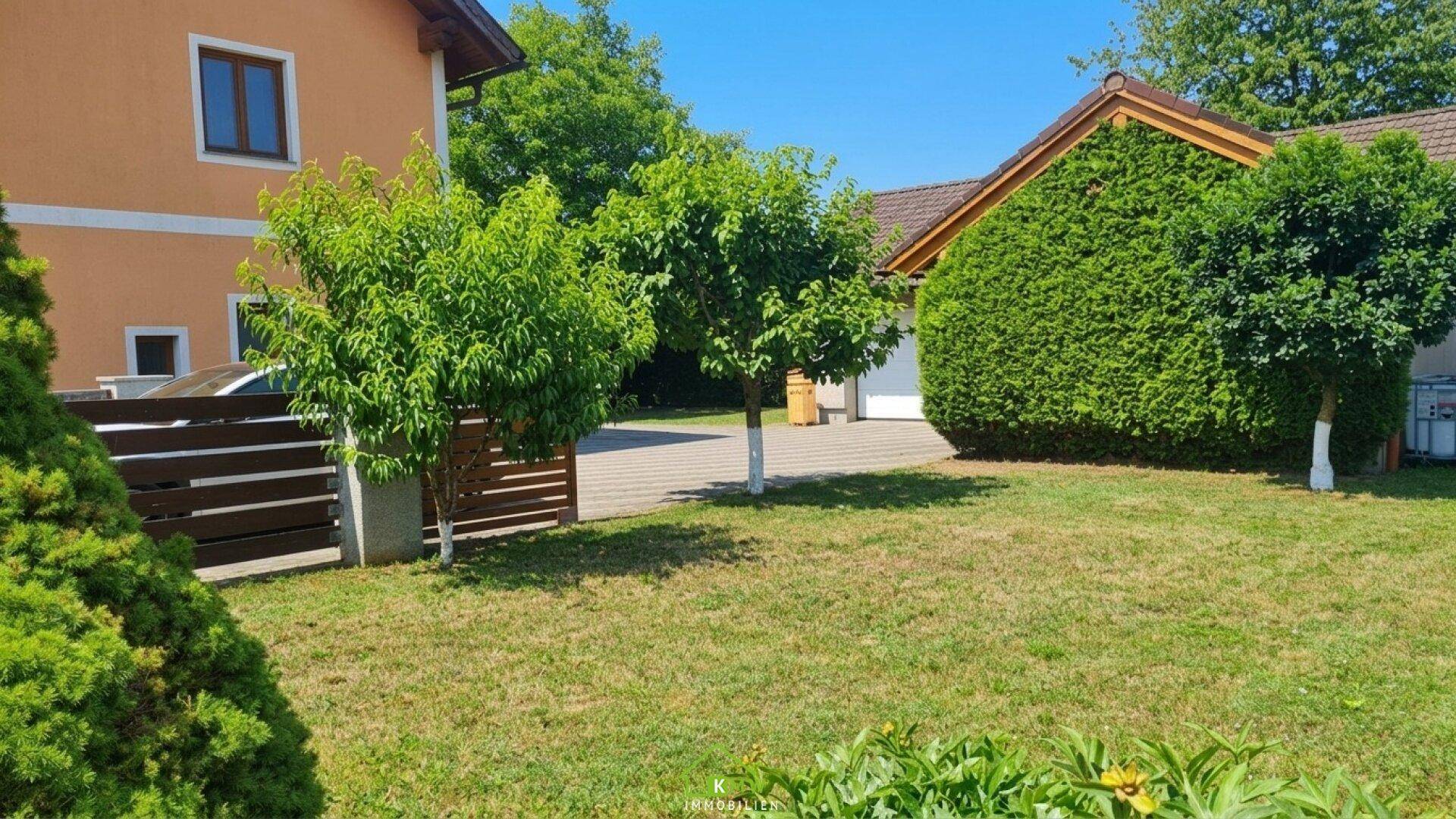 Grüner Gartenbereich mit Bäumen und gepflasterter Fläche vor einem Nebengebäude.