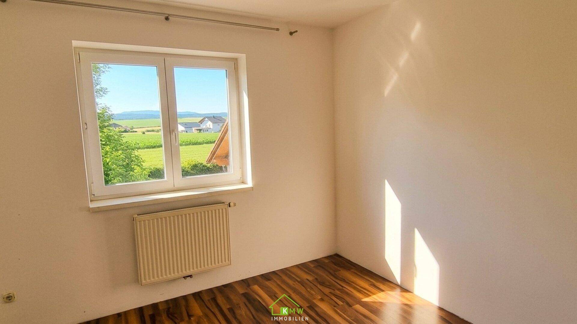 Schlafzimmer mit Laminatboden, weißer Wand und Fenster mit Blick ins Grüne.