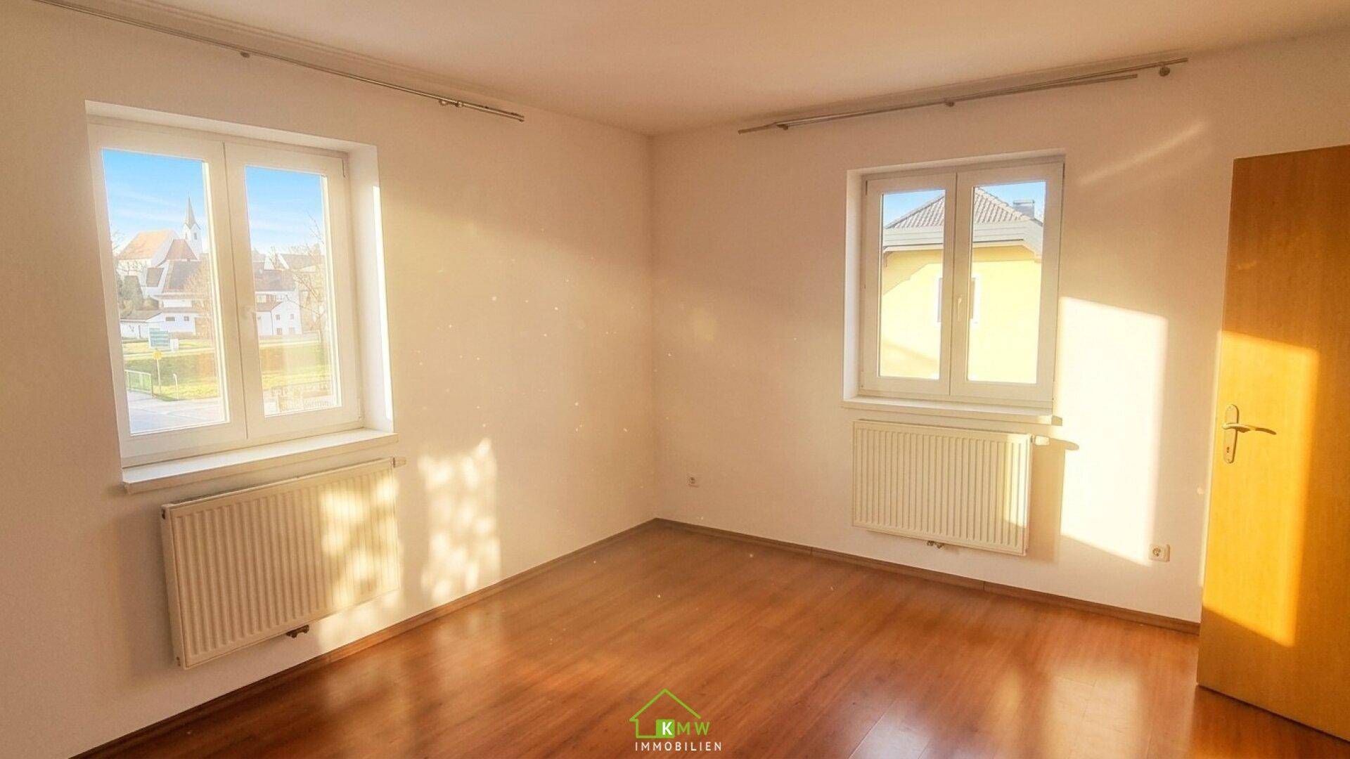 Sonniges Schlafzimmer mit Laminatboden, weißen Wänden und zwei Fenstern.