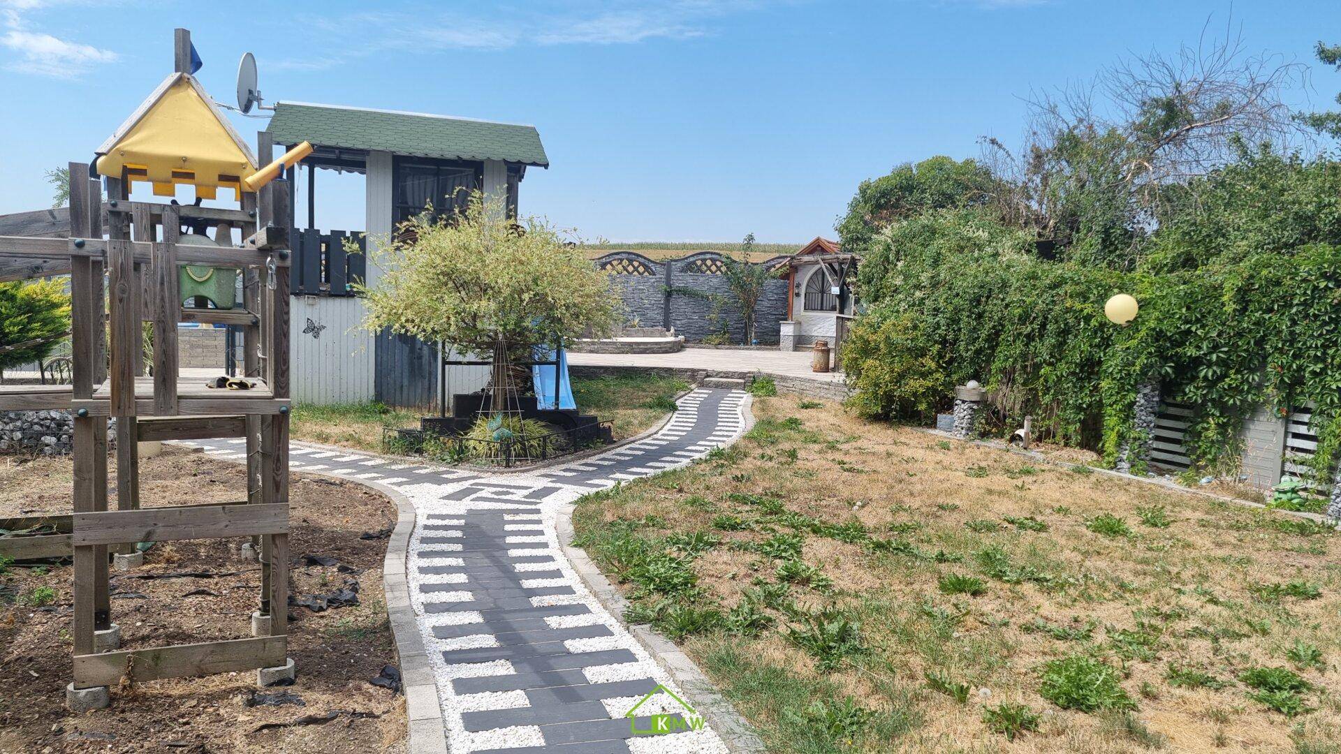 Großer Gartenbereich mit Spielturm, gepflasterten Wegen und viel Grünfläche unter blauem Himmel.
